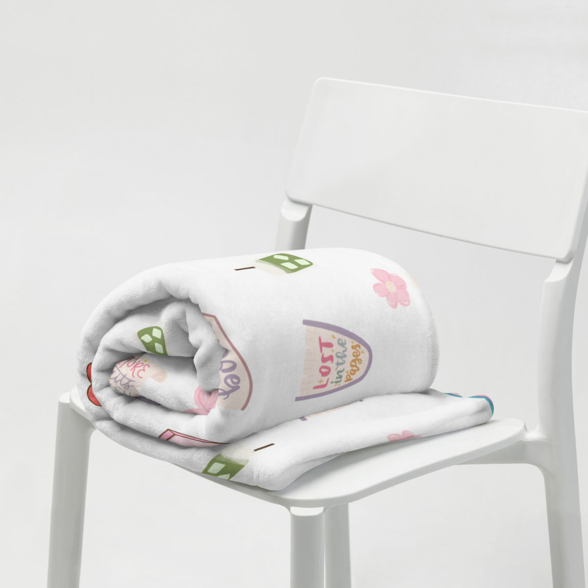 A white throw blanket with bookish illustrations is folded on a white chair.
