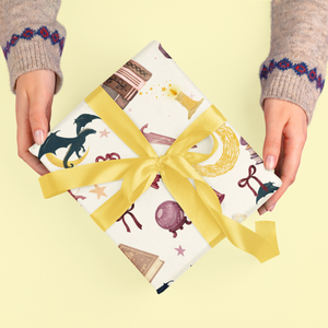 Hands hold a gift wrapped in paper with dragons, books, and stars, tied with a yellow ribbon.