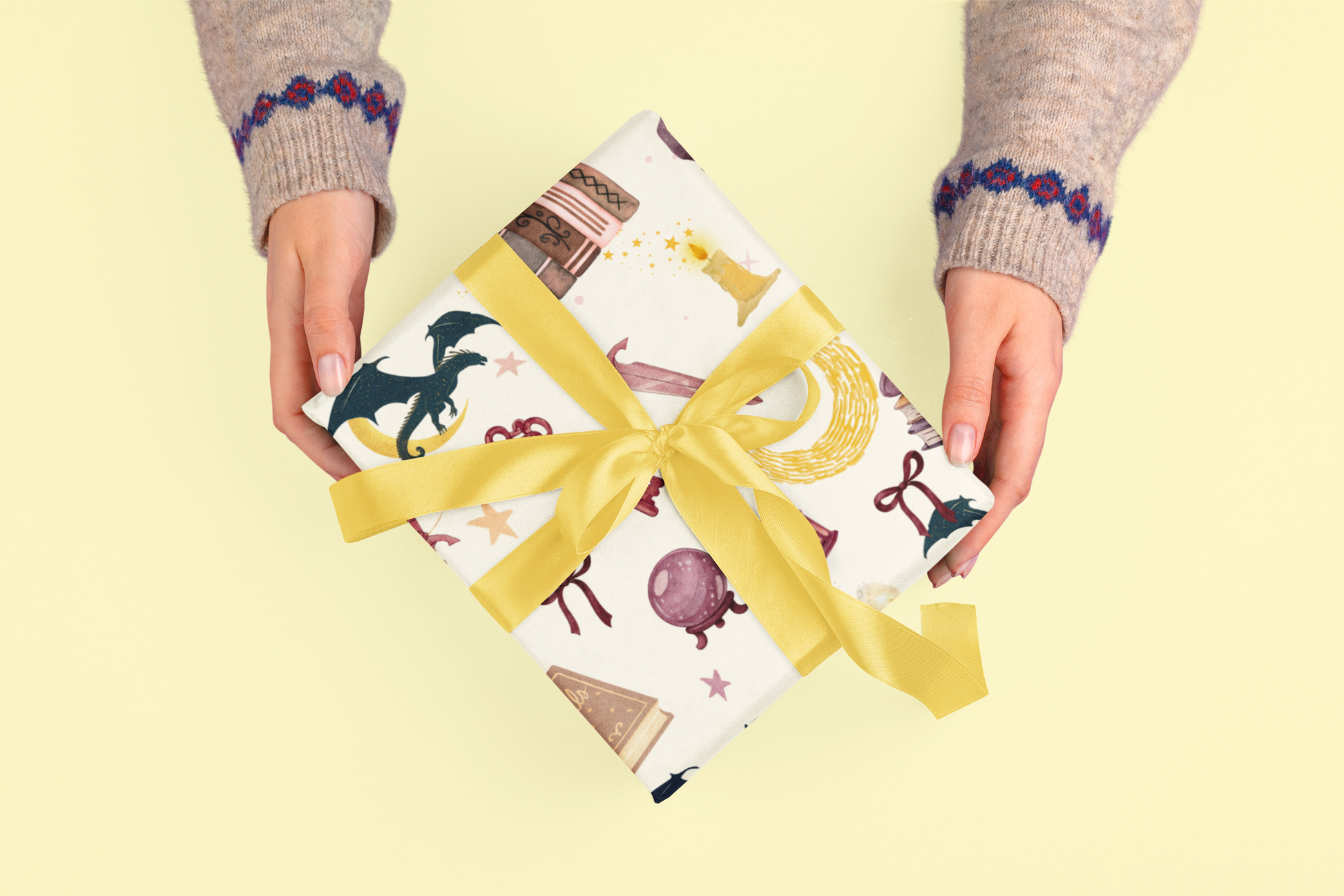 Hands hold a gift wrapped in paper with dragons, books, and stars, tied with a yellow ribbon.
