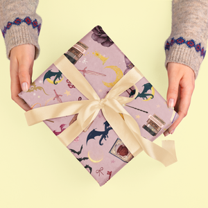 Hands hold a gift wrapped in purple paper with fantasy illustrations and a gold ribbon.